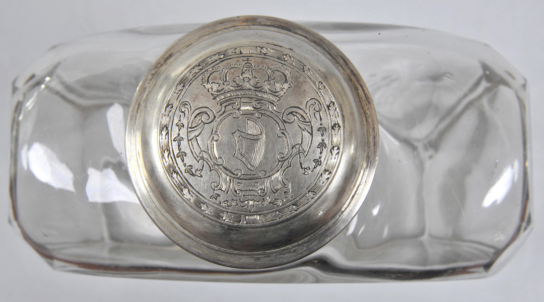 ujęcie z góry - zakrętka z grawerowanym herbem Janina. Jest on umieszczony w ukoronowanym, symetrycznym kartuszu z motywami wolutowymi i grirlandami z drobnych trójlistków. Przy krawędzi zgodnie z obwodem zakrętki, biegnie ozdobna wstęga.