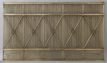 Ujęcie z przodu. Szeroki, metalowy obiekt. Krata stoi na metalowej listwie. W dolnej części widoczna ozdobna krata o eliptycznych otworach. Powyżej, aż do górnej krawędzi, przechodzi kilkadziesiąt cienkich prętów. Przez całą wysokość przechodzi również 6 grubszych prętów w równych odstępach. Między nimi w środku ukośne pręty tworzące znaki „X”. W miejscu ich przecięcia dekoracje w formie kwiatów. Na ich końcach – w formie liści kwiatu. Krata w kolorze złotym.