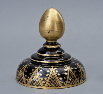 Strona pierwsza, ujęcie z boku. Pokrywa kopulasta. Na jej środku znajdują się trzy okrągłe, nieco wypukłe płytki. Położone są jedna na drugiej, każda z nich mniejsza od poprzedniej. Pokrywa zwieńczona jest jajowatą, złotą sterczyną na krótkiej szyjce. Na ściance kopuły znajdują się złocone dekoracje z motywem liści, pędów, gwiazd i linii, układające się w trójkąty.