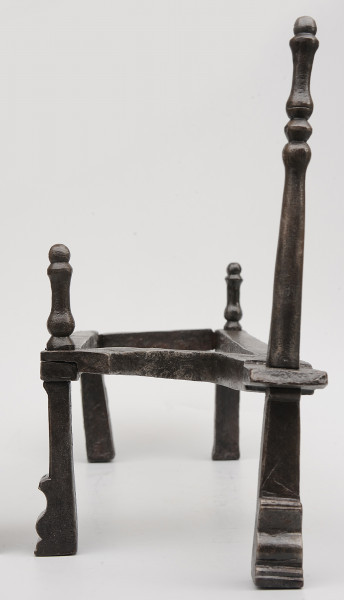 Strona pierwsza. Widok z przodu. Podstawka na opał wykonana z pręta, układającego się w figurę trapezu. Wsparty na czterech nogach. Nad nogą po prawej z przodu wysoka sterczyna. Dwie niższe sterczyny nad prawą nogą z tyłu i lewą nogą z przodu. Noga pod wyższą sterczyną wysunięta bardziej do przodu.