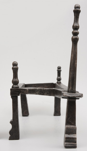 Strona pierwsza. Widok z przodu. Podstawka na opał wykonana z pręta, układającego się w figurę trapezu. Wsparty na czterech nogach. Nad nogą po prawej z przodu wysoka sterczyna. Dwie niższe sterczyny nad prawą nogą z tyłu i lewą nogą z przodu. Noga pod wyższą sterczyną wysunięta bardziej do przodu.