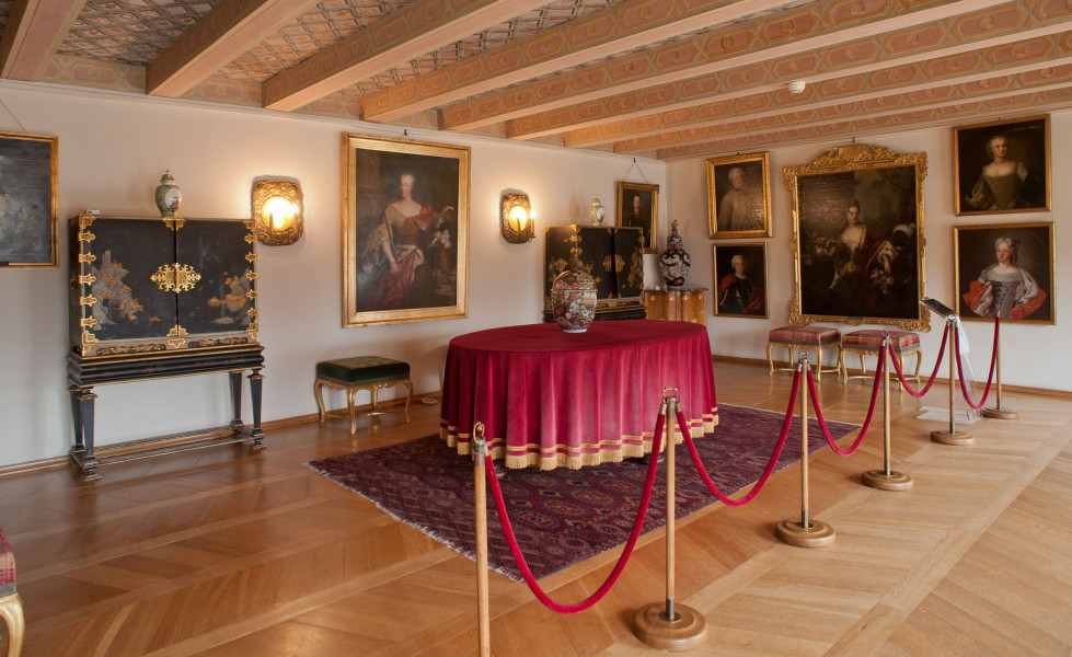 inwentaryzacja wystroju wnętrz, ściana północna, od góry belkowany sufit, na drugim planie ściana z wiszącymi obrazami, po lewej komoda w stylu chińskim na środku stół przykryty czerwonym obrusem.