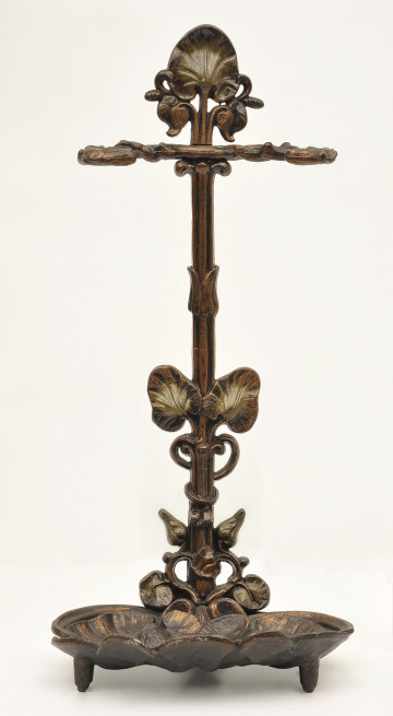 Ujęcie z przodu. Na dole zdobiona podstawka w formie nakładających się liści. Podstawka wsparta na widocznych dwóch nóżkach. Z tyłu od podstawki wychodzi pionowy trzpień oplatany dekoracjami w motywie liści. Na górze podpórka na przybory. Powyżej niej trzpień wieńczy płaska sterczyna.