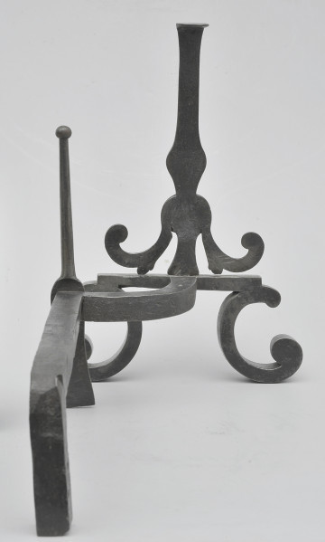 Ujęcie z tyłu. Na pierwszy planie widoczny metalowy, poziomy pręt, łączący się z smukłym szpikulcem. Przy szpikulcu pręt wygięty w prawo. Pręt łączy się z przednią częścią, w miejscu metalowej płytki. Na niej osadzony pionowy trzpień. Poniżej zwężenia na trzpieniu odchodzą dwa wygięte ramiona. Od płytki odchodzą wygięte stopki. Obiekt w całości czarny, lekko błyszczący.