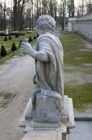 Ujęcie od strony drugiej. Bok rzeźby. Kolor szary. Postać mężczyzny siedzącego bokiem na niskim postumencie. Tułów skierowany w lewo. Głowa zwrócona do tyłu, lekko w lewo. Twarz jest niewidoczna. Przez lewe ramię przewieszona luźno tkanina opadająca na postument, zasłaniająca lewy bok postaci. W wyciągniętej lewej dłoni trzyma cylindryczny przedmiot. Włosy krótkie, zdobione liściem laurowym. W tle widoczny taras dolny Pałacu.