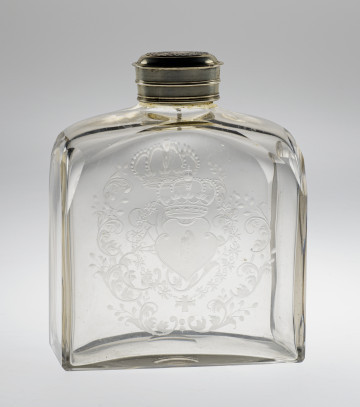 Ujęcie 3/4, z przodu, lekko z boku. Butla płaska, bezbarwna, o dwóch ściankach szerokich i dwóch wąskich. Ścianka z przodu szeroka. Widoczny wygrawerowany na niej herb Janina – wygięta tarcza skierowana w prawo. Herb otoczony zdobieniami. Przez szkło widoczne dekoracje na przeciwległej ściance. Ścianki na szczycie łagodnie się zwężają do srebrnego obramienia szyjki. Na butlę nałożona jest nakrętka.