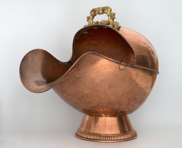 Ujęcie 3/4. Widok na przód i bok. Obiekt umieszczony na stopce o kształcie ściętego stożka. Pojemnik ma kształty zaokrąglone, z szerokim otworem o mocno wygiętej dolnej krawędzi po lewej stronie. Pojemnik i podstawa wykonane z miedzi, o kolorze jasnobrązowym. Na górze, przy krawędzi, widoczna rączka w kolorze złotym.