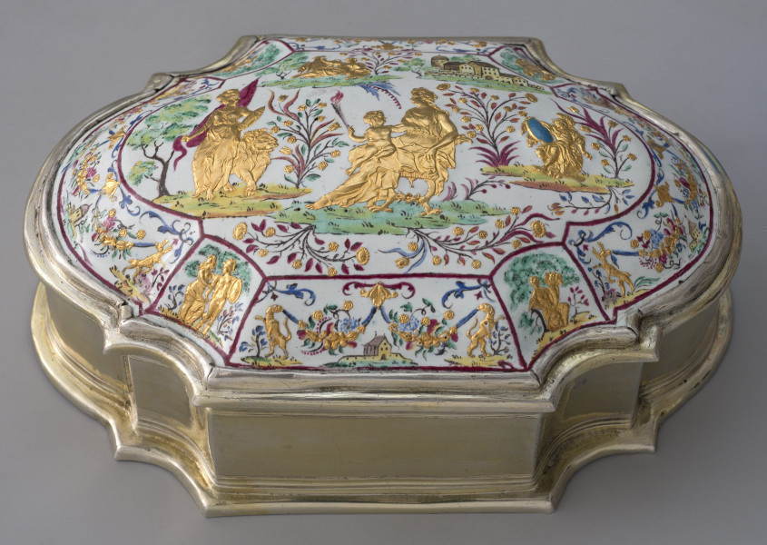 ujęcie lekko z góry, wieczko wypukłe bogato zdobione, na białym tle podzielone czerwoną cienką ramką na mniejsze sceny na skośnych elementach i scenę główną w centralnej części. Boczne sceny rodzajowe, leżąca po środku psy trzymające girlandy z kwiatów z zabudowaniami w tle, po prawej i lewej sceny rodzajowe złote postacie,  po bokach jelenie trzymające girlandy. Duża scena w centralnej części wieka, pejzaż cztery złote postacie, z lewej kobieta ze sztandarem i lwem po środku kobieta siedząca z dzieckiem na kolanach po prawej postać w zbroi z niebieską tarczą, w tle zabudowania. Bok puzderka złoty o zaokrąglonych węższych bokach i półokrągłymi wcięciami.