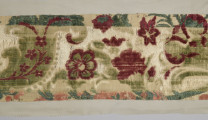 Pas tkaniny ułożony poziomo – zbliżenie na fragment. Widoczne motywy roślinne: czerwone kwiaty poprzeplatane zielonymi liśćmi na beżowym tle. W centralnej części szeroki pas tkaniny jest odbarwiony, liście są jaśniejsze, w tle jaśniejsze przeszycia. Wąski pas na dole również odbarwiony, motywy kwiatowe wpadają w pomarańcz. bryt Wil.Wn.126/1 - fragment, awers; ściana północno-wschodnia