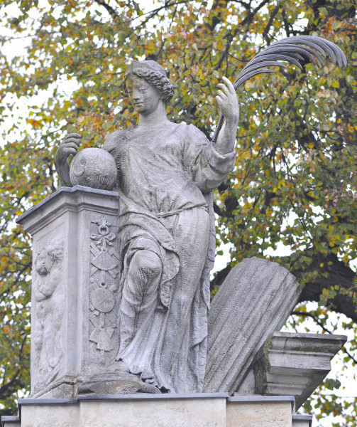 Pax - rzeźba na filarze południowym Bramy Głównej. Ujęcie z przodu, lekko z dołu. Postać kobiety oparta łokciem i biodrem o filar po lewej stronie. Kobieta ubrana w długą tunikę. Przy prawej dłoni, na filarze, widoczna kula. W lewej dłoni kobieta trzyma długie pióro. Twarz skierowana lekko w lewo i w dół. Po prawej, za postacią, widoczna przechylony nieco w lewo fragment kolumny. W tle gałęzie drzew.