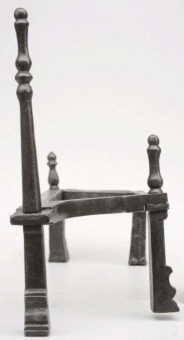 Strona pierwsza. Widok z przodu. Podstawka na opał wykonana z pręta, układającego się w figurę trapezu. Wsparty na czterech nogach. Nad nogą po lewej z przodu wysoka sterczyna. Dwie niższe sterczyny nad lewą nogą z tyłu i prawą nogą z przodu. Noga pod wyższą sterczyną wysunięta bardziej do przodu.