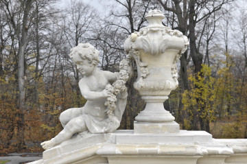 ujęcie od strony pierwszej. Putto z lewej, wazon z prawej, putto ustawione na pochyłej balustradzie schodów, wazon na poziomej. Putto ma tułów silnie skręcony w lewą stronę. W rękach wyciągniętych w lewo trzyma kwiatową girlandę. Lewe udo przykryte draperią przewieszoną przez lewe ramię. Głowa zwrócona w prawo. Obie figury w kolorze szarobeżowym. Obok waza o kielichowatym kształcie dekorowana girlandami kwiatów i stylizowanymi liśćmi. W tle drzewa parkowe.