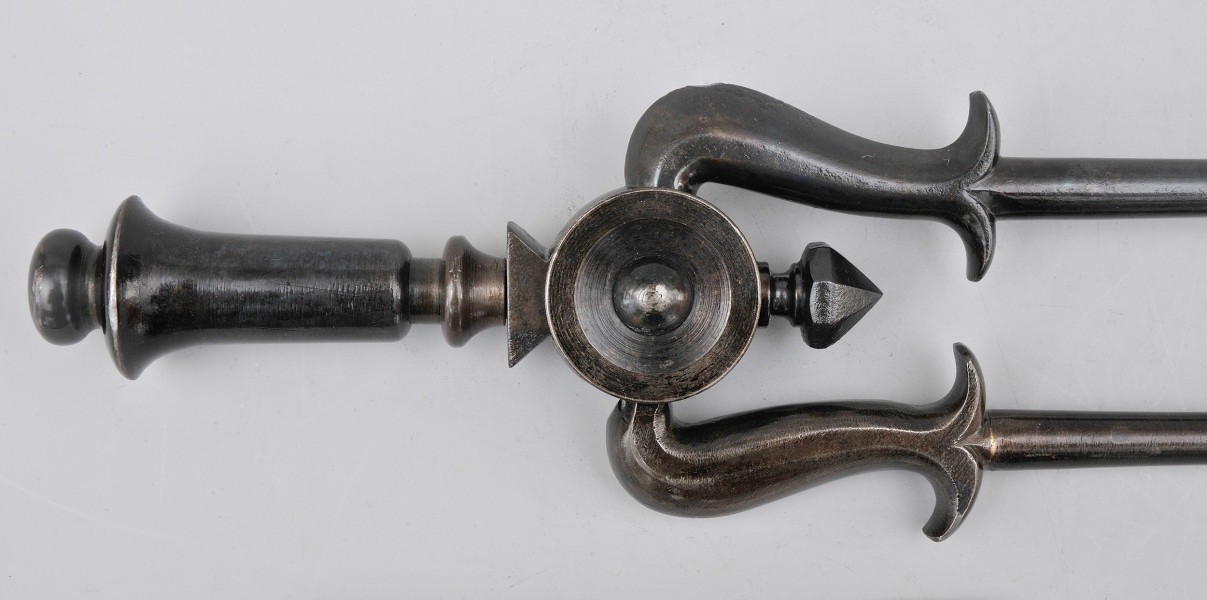 Fragment – uchwyt. Szczypce ułożone poziomo, z ramionami skierowanymi w prawo. Dzwonowata rączka zwieńczona kulką. Rączka łączy się z przegubem, ozdobionym w środku wypukłością. Na prawo od niej łączy się z zdobieniem w formie ostrosłupa. Od przegubu w górę i w dół odchodzą szczypce, które są wygięte w prawo. Widoczne wywinięte wąsy na ramionach szczypiec. Pozostała cześć obiektu niewidoczna w kadrze. Szczypce czarne, błyszczące.