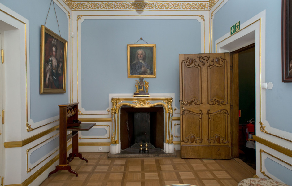 ściana południowa, centralnie kominek zdobiony złotymi płomieniami oraz elementami roślinnymi, po prawej otwarte drewniane drzwi, ściany błękitno białe ze złoceniami w formie pozłacanych listewek z narożami z motywami roślinnymi wykończone u góry szerokim złotym fryzem z motywami roślinnymi, nad kominkiem obraz, po lewej sekretarzyk i obraz wiszący na ścianie.