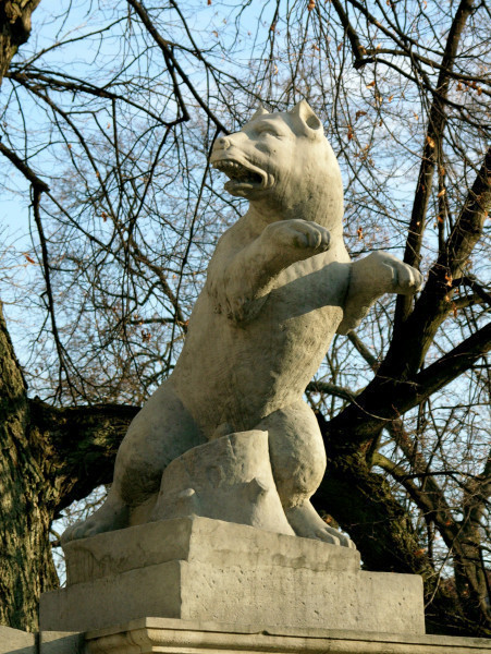 Pies - rzeźba na przyporze południowej Bramy Głównej. Widok lekko z lewej i od dołu. Pies stojący na dwóch tylnych łapach. Dwie przednie łapy wyciągnięte przed siebie. Łeb psa skierowany w lewo, pysk rozwarty. Między tylnymi łapami widoczny pień drzewa. Kamienna figura ma jasny, piaskowy kolor, za nią widać konary bezlistnego drzewa.