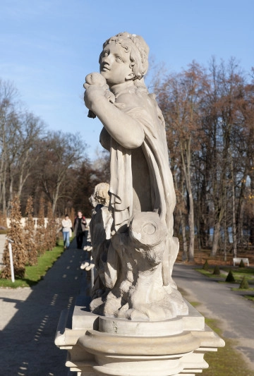 Ujęcie od strony drugiej, bok rzeźby. Kolor szaro-beżowy. Postać kobiety siedzi na konarze drzewa. Ustawiona bokiem do widza. Głowa skierowana lekko w lewo i w górę. W lewej dłoni, zgiętej w łokciu i uniesionej do góry, trzyma jabłko. Przez lewe ramię przewieszona jest tkanina. W tle widoczne tarasy za Pałacem i drzewa parkowe.