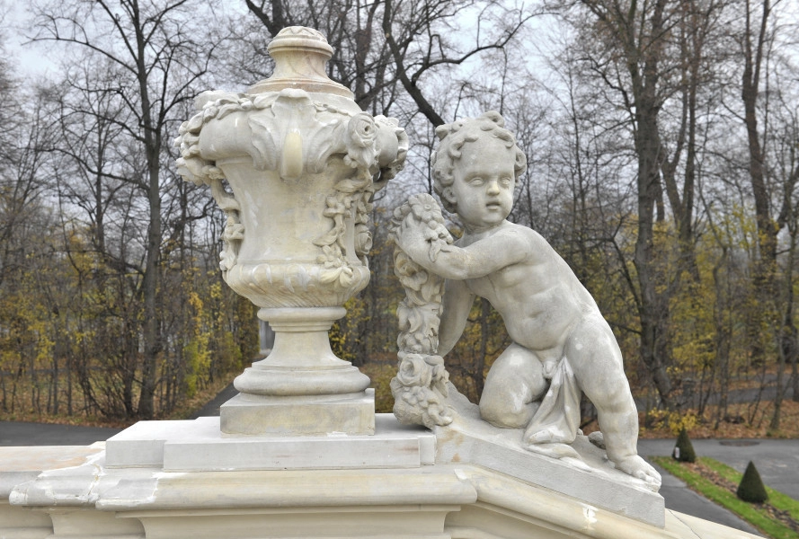 ujęcie od strony pierwszej. Putto z prawej, wazon z lewej. Putto ustawione na pochyłej balustradzie schodów, wazon na poziomej. Putto oparte na prawym kolanie, lewa noga wyprostowana. Prawa ręka wyprostowana wsparta na podstawie, w lewej ręce girlanda, tułów silnie skręcony w prawą stronę. Chłopiec zwrócony twarzą w stronę widza. Obok waza o kielichowatym kształcie dekorowana girlandami kwiatów i stylizowanymi liśćmi. Obie figury w kolorze beżowym. W tle drzewa parkowe.