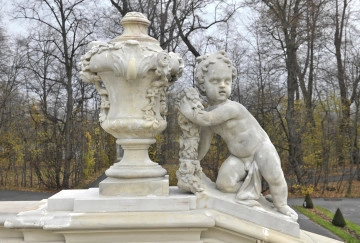 ujęcie od strony pierwszej. Putto z prawej, wazon z lewej. Putto ustawione na pochyłej balustradzie schodów, wazon na poziomej. Putto oparte na prawym kolanie, lewa noga wyprostowana. Prawa ręka wyprostowana wsparta na podstawie, w lewej ręce girlanda, tułów silnie skręcony w prawą stronę. Chłopiec zwrócony twarzą w stronę widza. Obok waza o kielichowatym kształcie dekorowana girlandami kwiatów i stylizowanymi liśćmi. Obie figury w kolorze beżowym. W tle drzewa parkowe.