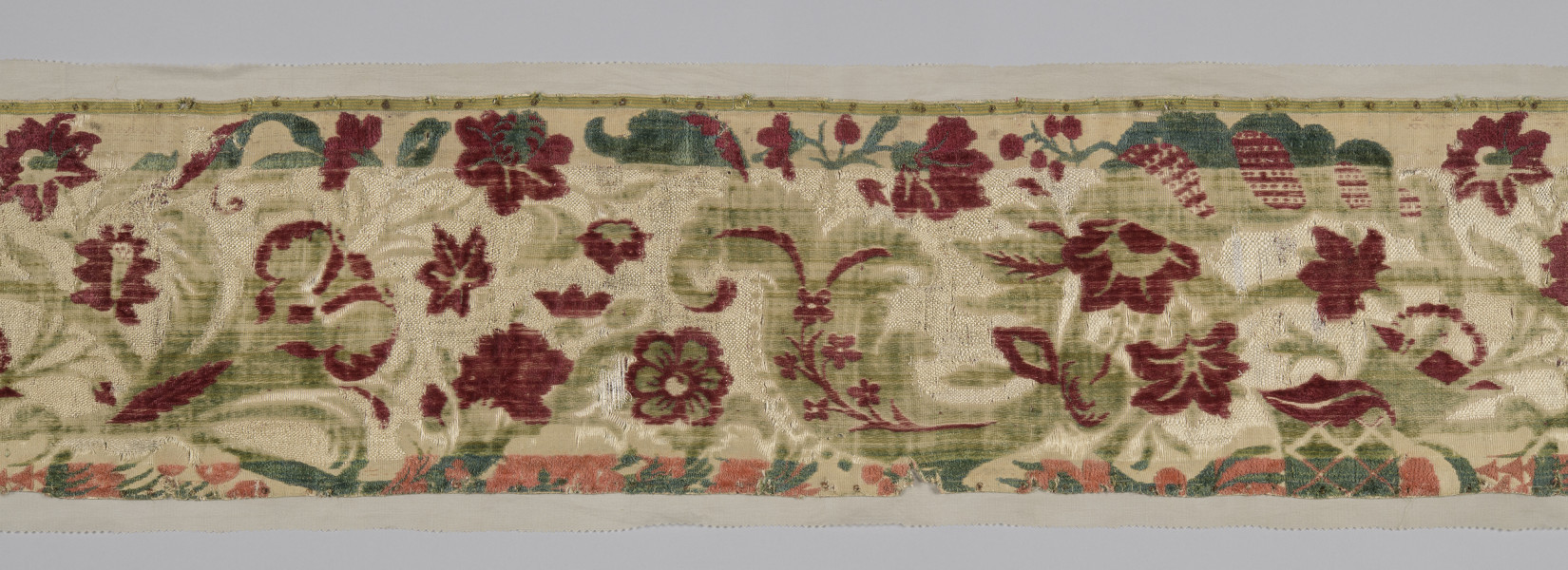 Pas tkaniny ułożony poziomo. Widoczne motywy roślinne: czerwone kwiaty poprzeplatane zielonymi liśćmi na beżowym tle. W centralnej części szeroki pas tkaniny jest odbarwiony, liście są jaśniejsze, w tle jaśniejsze przeszycia. Wąski pas na dole również odbarwiony, motywy kwiatowe wpadają w pomarańcz. bryt Wil.Wn.125/1 - fragment, awers; ściana południowo-wschodnia