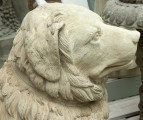 zbliżenie na pysk psa. Pies zwrócony pyskiem w prawo. Rasą podobny do golden retrievera: puszyste futro, oklapnięte uszy, ma obrożę. Figura w kolorze beżowym. W tle inne rzeźby ustawione na drewnianych paletach. 