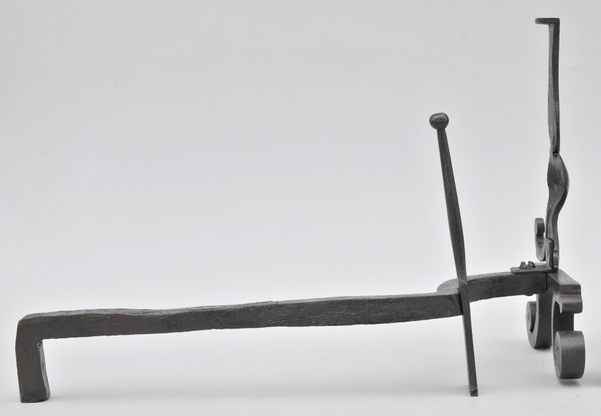 Ujęcie ze wspornikiem na polano. Widok z boku, z przodem po prawej stronie. W większości pręt prosty, bliżej przodu wygięty w lewo. W miejscu wygięcia smukły szpikulec. Pręt łączy się z przednią częścią obiektu przy metalowej płytce. Powyżej niej pionowy trzpień. Poniżej zwężenia na trzpieniu odchodzi w dół wygięte ramię w stronę drugiego planu. Po drugiej stronie widoczny ubytek. Od płytki odchodzą wygięte stopki. Obiekt w całości czarny, lekko błyszczący.