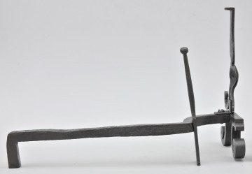Ujęcie ze wspornikiem na polano. Widok z boku, z przodem po prawej stronie. W większości pręt prosty, bliżej przodu wygięty w lewo. W miejscu wygięcia smukły szpikulec. Pręt łączy się z przednią częścią obiektu przy metalowej płytce. Powyżej niej pionowy trzpień. Poniżej zwężenia na trzpieniu odchodzi w dół wygięte ramię w stronę drugiego planu. Po drugiej stronie widoczny ubytek. Od płytki odchodzą wygięte stopki. Obiekt w całości czarny, lekko błyszczący.