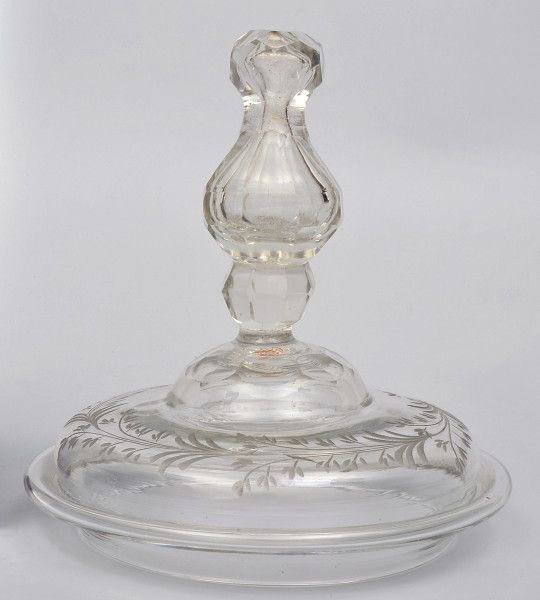 strona pierwsza.  Na górze, ukazanej z boku okrągłej pokrywy, umieszczony wysoki, profilowany i fasetowany uchwyt. Powierzchnia pokrywy jest dekorowana motywem roślinnym.  Na górze zgodnie z obwodem pokrywy biegnie, matowo rytowany otok z gałązek o drobnych listkach.