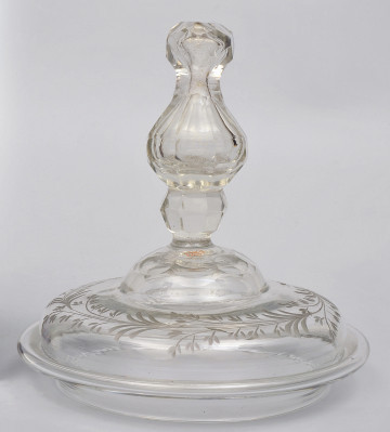 strona pierwsza.  Na górze, ukazanej z boku okrągłej pokrywy, umieszczony wysoki, profilowany i fasetowany uchwyt. Powierzchnia pokrywy jest dekorowana motywem roślinnym.  Na górze zgodnie z obwodem pokrywy biegnie, matowo rytowany otok z gałązek o drobnych listkach.