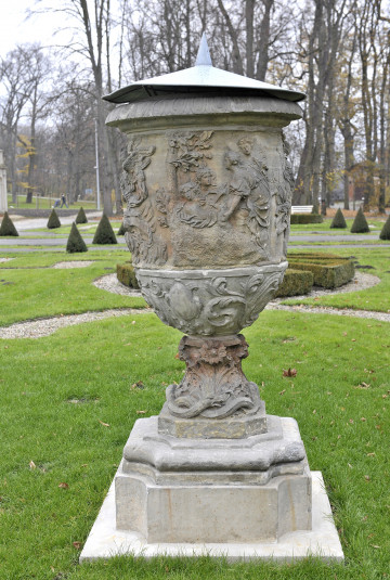 ujęcie od strony pierwszej, widok z boku, wazon przykryty metalową nakrywką z ostrym trzpieniem, czasza wazonu ozdobiona sceną mitologiczną postacie kobiece przy drzewie, dół i noga wazonu ozdobiona wzorami kwiatowymi, u podstawy dekoracja przestawiająca wijące się węże, wazon stoi na cokole wśród trawy w tle stożki cisów oraz drzewa bez liści.