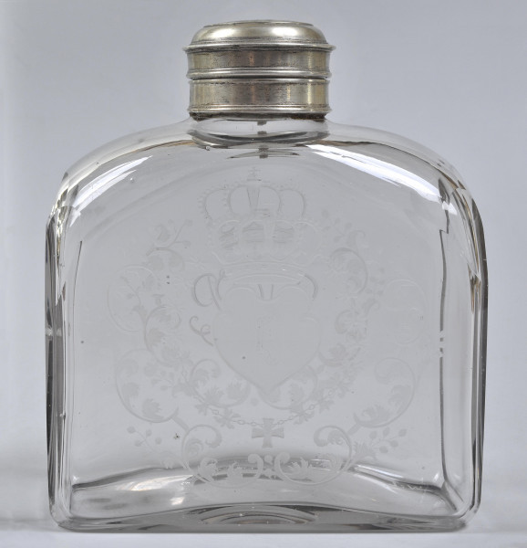 Strona trzecia, widok na szeroką, płaską ścianę. Butelka bezbarwna. Ledwo widoczny wygrawerowany herb Janina – wygięta tarcza skierowana w prawo. Herb otoczony zdobieniami. Ścianka na szczycie łagodnie się zwęża do srebrnego obramienia szyjki. Na butlę nałożona jest nakrętka. Przez szkło widoczne dekoracje na przeciwległej ściance.