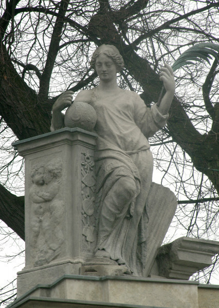 Pax - rzeźba na filarze południowym Bramy Głównej. Ujęcie lekko z lewej i z dołu. Postać kobiety w diademie na głowie, odziana w długą, powłóczystą szatę, przewiązaną na wysokości bioder sznurem,  oparta łokciem i biodrem o filar po lewej stronie. Przy prawej dłoni, na filarze, widoczna kula. W lewej dłoni kobieta trzyma długie pióro. Twarz skierowana w kierunku widza, lekko w dół. W tle gałęzie drzew.