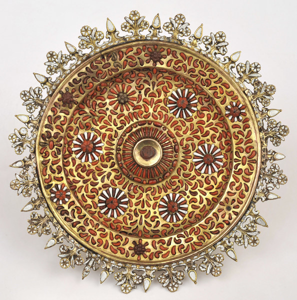 ujęcie z góry. Okrągła złota patera bogato zdobiona białą emalią i czerwonym koralem w kształcie płatków kwiatów i w biało-czerwone wzory kwiatowe, brzeg patery ażurowy, zdobiony bukietami kwiatów.