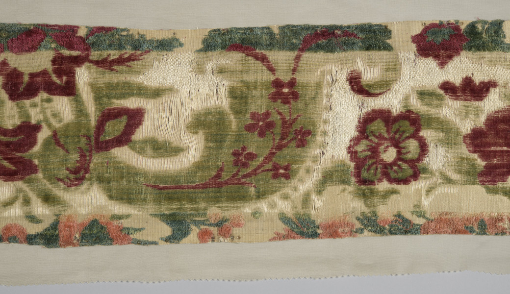 Pas tkaniny ułożony poziomo – zbliżenie na fragment. Widoczne motywy roślinne: czerwone kwiaty poprzeplatane zielonymi liśćmi na beżowym tle. W centralnej części szeroki pas tkaniny jest odbarwiony, liście są jaśniejsze, w tle jaśniejsze przeszycia. Wąski pas na dole również odbarwiony, motywy kwiatowe wpadają w pomarańcz. bryt Wil.Wn.126/1 - fragment, awers; ściana północno-wschodnia