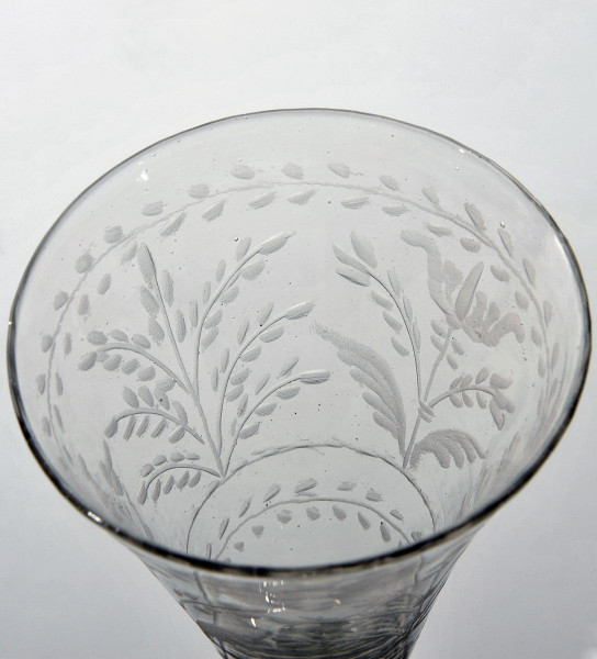 ujęcie lekko z góry - fragment wnętrza kieliszka z matowo rytowaną dekoracja liściastą.. W pasie dekoracji ograniczonej przez dwie linie z ukośnie umieszczonymi listkami, pionowo ukazane dwie rośliny. Po lewej roślina z liściastymi gałązkami, po prawej kwiat podobny do tulipana.
