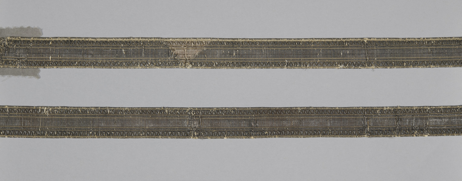 Dwie tasiemki ułożone poziomo, równolegle do siebie. Obydwie wykonane według tego samego wzoru w kolorze ciemnobrązowym, wewnątrz cienkie linie ze złotej nitki: na krawędziach i dwie pary wewnątrz wstęgi, symetrycznie względem osi tasiemki. Na górnej tasiemce widoczne trójkątne odbarwienie w kolorze jasnobrązowym. galon Wil.Wn.127/7 - fragment, awers; ściana północna