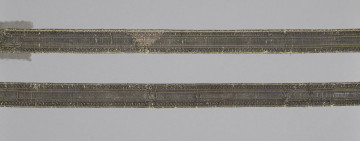 Dwie tasiemki ułożone poziomo, równolegle do siebie. Obydwie wykonane według tego samego wzoru w kolorze ciemnobrązowym, wewnątrz cienkie linie ze złotej nitki: na krawędziach i dwie pary wewnątrz wstęgi, symetrycznie względem osi tasiemki. Na górnej tasiemce widoczne trójkątne odbarwienie w kolorze jasnobrązowym. galon Wil.Wn.127/7 - fragment, awers; ściana północna