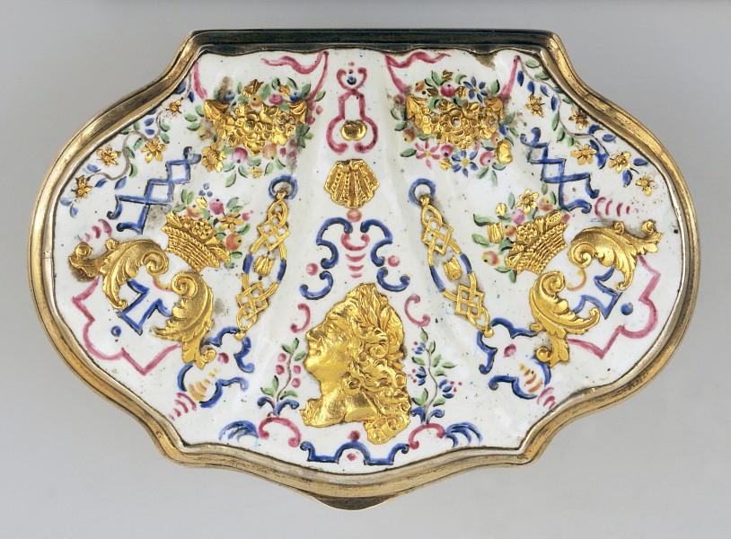 ujęcie z góry na pokrywkę w kształcie muszli na białym tle po środku złote popiersie, po bokach symetrycznie złote palmety oraz kosze z owocami i kwiatami. Ornamenty złote z niebieskimi i czerwonymi wzorami.