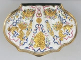 ujęcie z góry na pokrywkę w kształcie muszli na białym tle po środku złote popiersie, po bokach symetrycznie złote palmety oraz kosze z owocami i kwiatami. Ornamenty złote z niebieskimi i czerwonymi wzorami.