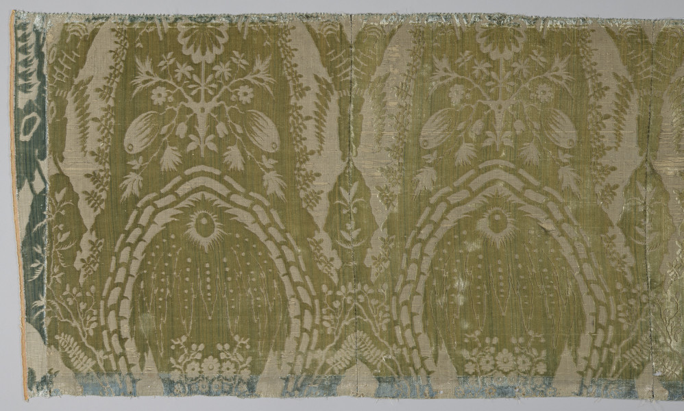 Pas tkaniny z motywami roślinnymi w kolorze żółto-zielonym. Na górze i na dole odbarwione na niebiesko pasy. Po lewej przyszyty jest materiał w kolorze niebieskim. Widoczne pionowe szwy. bryt Wil.Wn.133/3 - fragment, awers; ściana północna