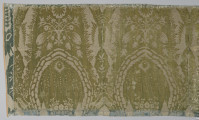Pas tkaniny z motywami roślinnymi w kolorze żółto-zielonym. Na górze i na dole odbarwione na niebiesko pasy. Po lewej przyszyty jest materiał w kolorze niebieskim. Widoczne pionowe szwy. bryt Wil.Wn.133/3 - fragment, awers; ściana północna