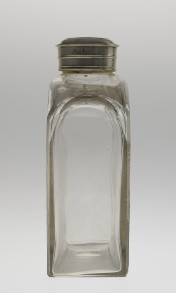 Lewy bok, widok na całość butli. Ścianka wąska, wysoka i płaska, bezbarwna. Na szczycie łagodnie się zwęża do srebrnego obramienia szyjki. Na butlę nałożona jest nakrętka. Na jej boku jest wygrawerowany napis „N.1.”.