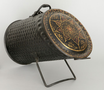 Ujęcie 3/4. Pojemnik widoczny od przodu i lekko z boku. Nachylony lekko w górę, oparty o tylną krawędź i podpórkę. Kształt cylindryczny, ścięty po skosie. Po prawej stronie ujęcia widać zdobioną, zamkniętą klapę pojemnika. Ścianki pojemnika są w kolorze czarnym i pokryte uwypukleniami.