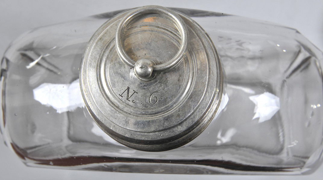 Ujęcie z góry - zatyczka-korek. Zatyczka okrągła, srebrna. Na wierzchu grawerowane koncentryczne okręgi, wewnątrz nich wygrawerowany napis „N.6.”. W górnej części kadru uchwyt w formie niewielkiej obręczy, przewleczony przez kulistą wypukłość na środku. Na drugim planie widoczna szeroka, płaska butla o płaskich i przezroczystych ściankach.