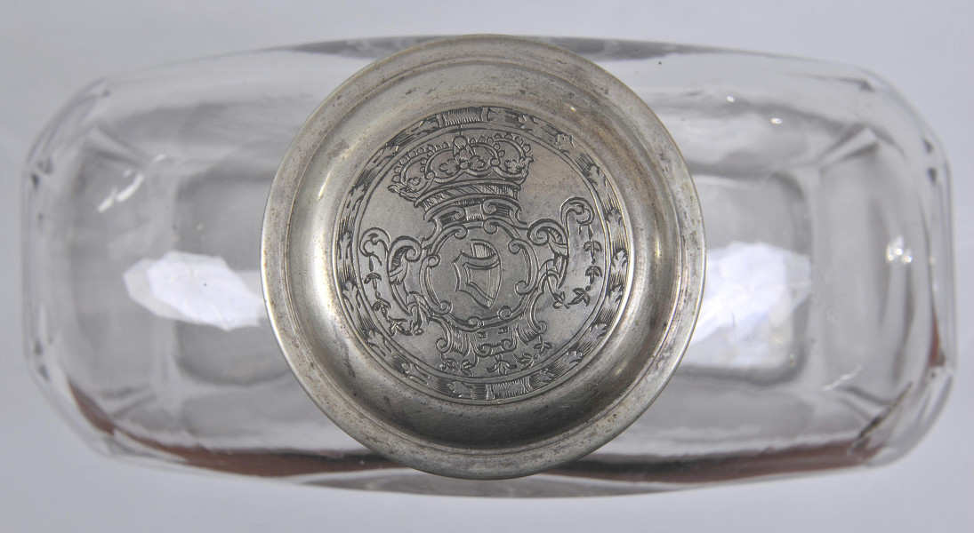 Ujęcie z góry - zakrętka z grawerowanym herbem Janina - wygiętą tarczą skierowaną w prawo. Nad herbem korona królewska, a całość otoczona wieńcem. Sama nakrętka przekręcona jest lekko w lewo. Na drugim planie widoczna szeroka, płaska butla o płaskich i przezroczystych ściankach.