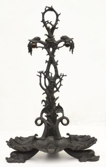 Ujęcie z przodu. Na dole zdobiona podstawka w formie potrójnej muszli. Pod podstawką widać dwie nóżki w postaci delfinów, których ogony wykrzywiają się w górę za podstawką. Między ogonami osadzony jest trzpień w formie rozwidlającej się gałęzi. U góry gałęzie tworzą podpórkę na przybory kominkowe. Powyżej niej sterczyna w formie pętli z gałęzi. Stojak w kolorze czarnym.