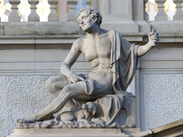 Ujęcie od strony pierwszej strony. Kolor rzeźby szaro-żółty. Postać mężczyzny siedzącego bokiem na niskim postumencie. Tułów skierowany w stronę widza. Nogi ułożone w lewo, prawa zgięta w kolanie. Głowa skierowana w prawo i lekko do góry. Postać naga, przez jego lewe ramię przewieszona jest luźno tkanina okrywająca lewy bok mężczyzny. Tkanina przechodzi za plecami na uda. Prawa ręka oparta na nogach, z wyciągniętym palcem wskazującym. Lewa ręka, wyciągnięta w prawą stronę, trzyma cylindryczny przedmiot. Włosy krótkie, zdobione liściem laurowym. Pod lewą nogą leży mały pies. W tle widoczna ściana tarasu, nieco wyżej widać też balustradę.