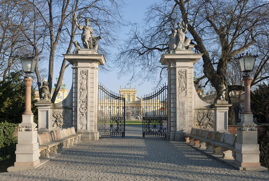 Widok na Bramę Główną od strony Przedpola, przęsła metalowe przymknięte. Brama w kolorze beżowym, o bogato zdobionej elewacji. Rzeźby mają kolor szary. Po lewej i prawej na przyporach rzeźby kolejno niedźwiedzia i psa. Łączące się niewielkim łukiem z wysokimi filarami. Na filarach widoczne rzeźby. Na lewym – Mars, a na prawym – Pax. W tle widoczny korpus Pałacu.