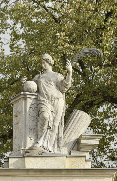 Pax - rzeźba na filarze południowym Bramy Głównej. Ujęcie z przodu, lekko z dołu. Postać kobiety oparta łokciem i biodrem o filar po lewej stronie. Kobieta ubrana w długą tunikę. Przy prawej dłoni, na filarze, widoczna kula. W lewej dłoni kobieta trzyma długie pióro. Twarz skierowana lekko w lewo i w dół. Po prawej, za postacią, widoczna przechylony nieco w lewo fragment kolumny. W tle gałęzie drzew.