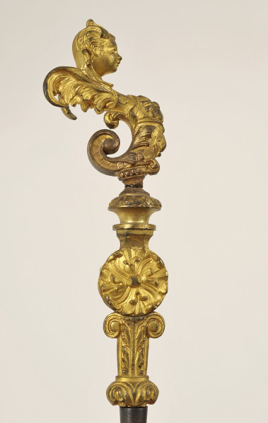 Detal rękojeści - strona prawa. Widok od boku. Rękojeść bogato zdobiona w kolorze złotym. Widoczne motywy liści, powyżej niej rozeta, a jeszcze wyżej mocno wygięta w lewo figurka hermy o dziecięcej głowie. Głowa skierowana w prawo.