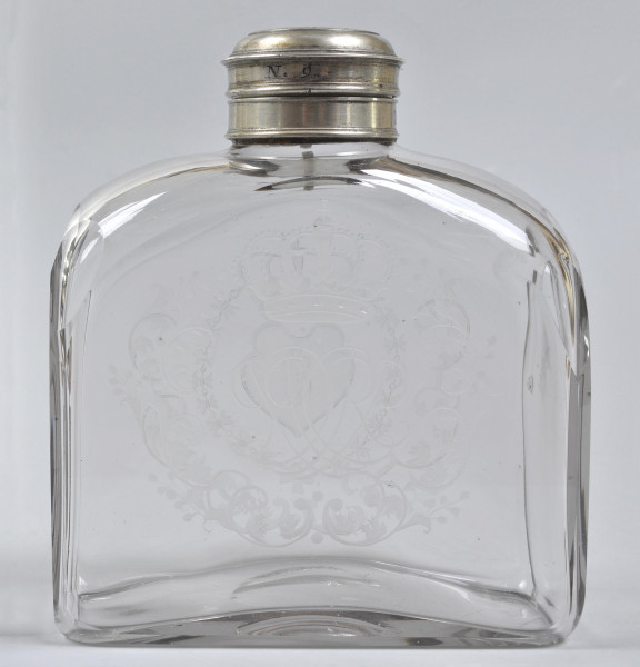 Strona pierwsza, widok na szeroką, płaską ścianę. Butelka bezbarwna. Ledwo widoczny wygrawerowany monogram „CRP”. Monogram otoczony zdobieniami. Ścianka na szczycie łagodnie się zwęża do srebrnego obramienia szyjki. Na butlę nałożona jest nakrętka. Na jej boku jest wygrawerowany napis „N.9.”. Przez szkło widoczne dekoracje na przeciwległej ściance.